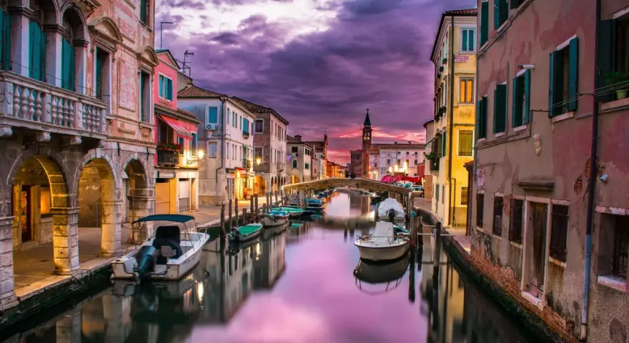Un canale di Venezia al tramonto, con barche ormeggiate e riflessi viola sull'acqua. Un'atmosfera magica che ricorda i piombi di Venezia.