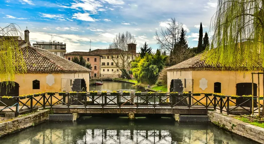 Ponte in legno su un canale con mulini ad acqua, case storiche e vegetazione rigogliosa, un'atmosfera serena che ricorda Portogruaro Caorle.