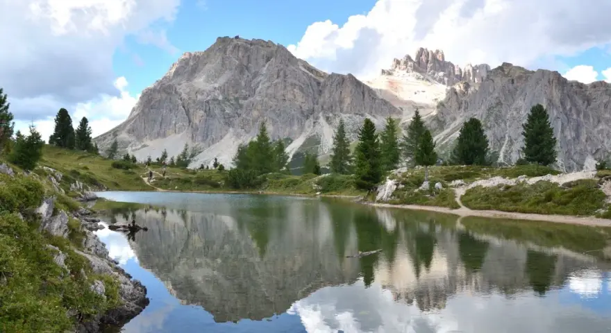 Riflessi perfetti delle montagne nel **lago di Limedes**, con sentiero e pini che incorniciano il paesaggio.