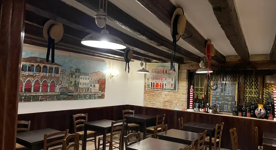 Interno di un **ristorante antico** con dipinti di **Venezia**, tavoli in legno e un calice di vino.