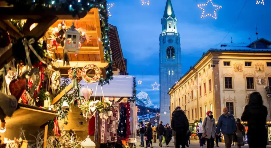 Mercatino di Natale a Cortina d'Ampezzo, con bancarelle addobbate, luci scintillanti e la torre del campanile che domina il cielo serale.