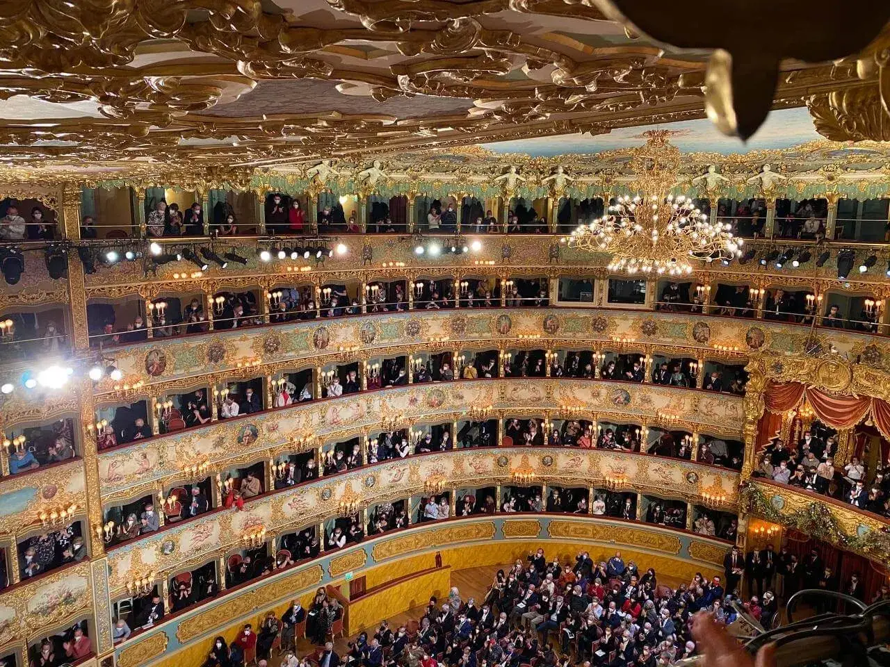 Interni sfarzosi del Teatro La Fenice di Venezia, con pubblico in maschera seduto nelle logge dorate sotto un lampadario scintillante.