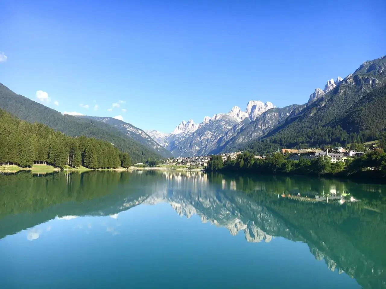Il lago Auronzo di Cadore riflette montagne maestose e un cielo azzurro. Un villaggio si affaccia sulle sue acque calme.