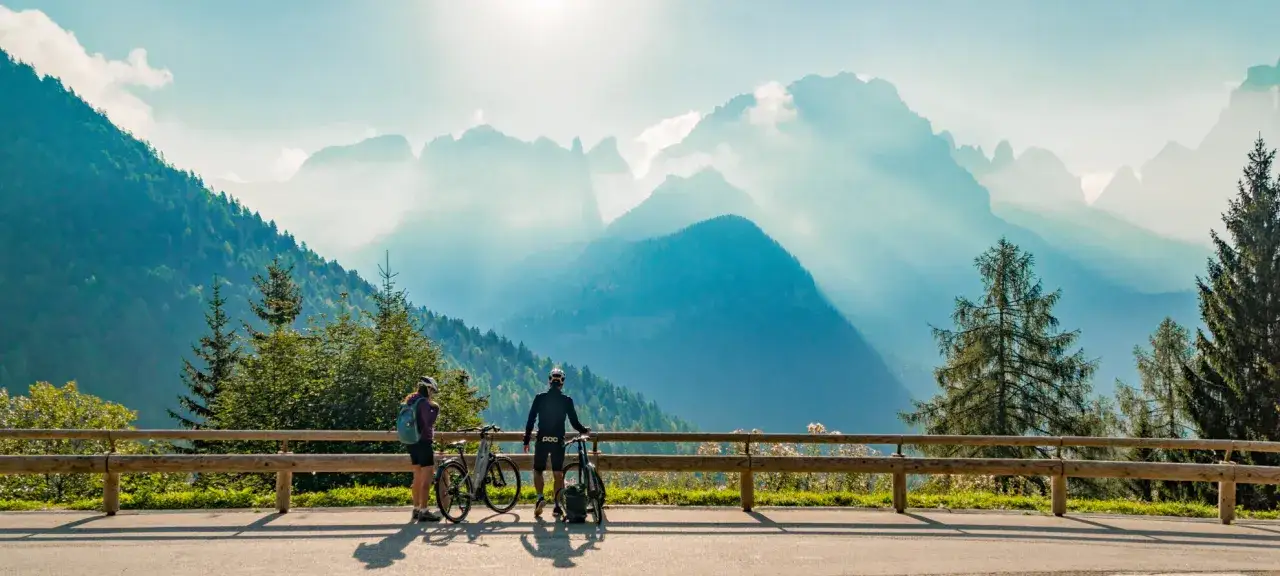 Coppia di ciclisti ammira il panorama mozzafiato delle ciclabili Dolomiti. Montagne azzurre e boschi verdi sotto un cielo limpido.