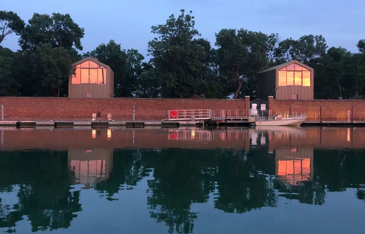 Riflessi dorati al tramonto su due case moderne con grandi finestre, un molo e una barca. Un angolo di Venezia segreta.