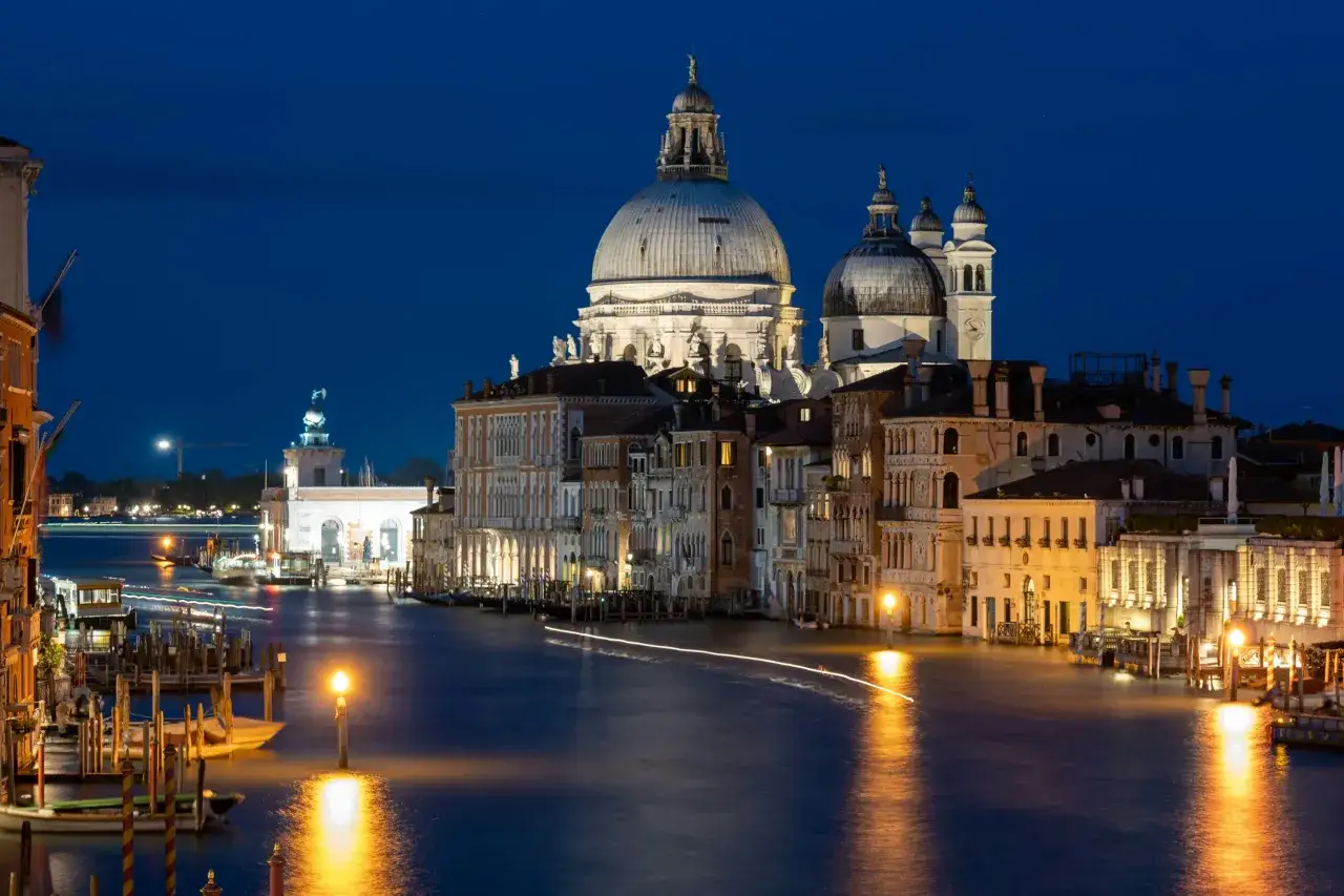 Venezia di notte, con la Basilica di Santa Maria della Salute illuminata e riflessa sull'acqua scura del Canal Grande, mentre le luci delle imbarcazioni creano scie luminose.