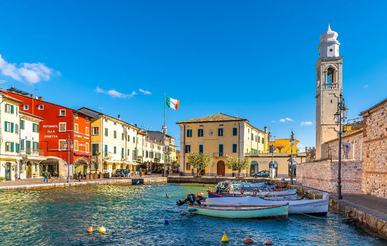 Un pittoresco porto con case colorate e un campanile, ideale per scoprire cosa vedere a Lazise in un giorno. Barche ormeggiate sull'acqua azzurra.