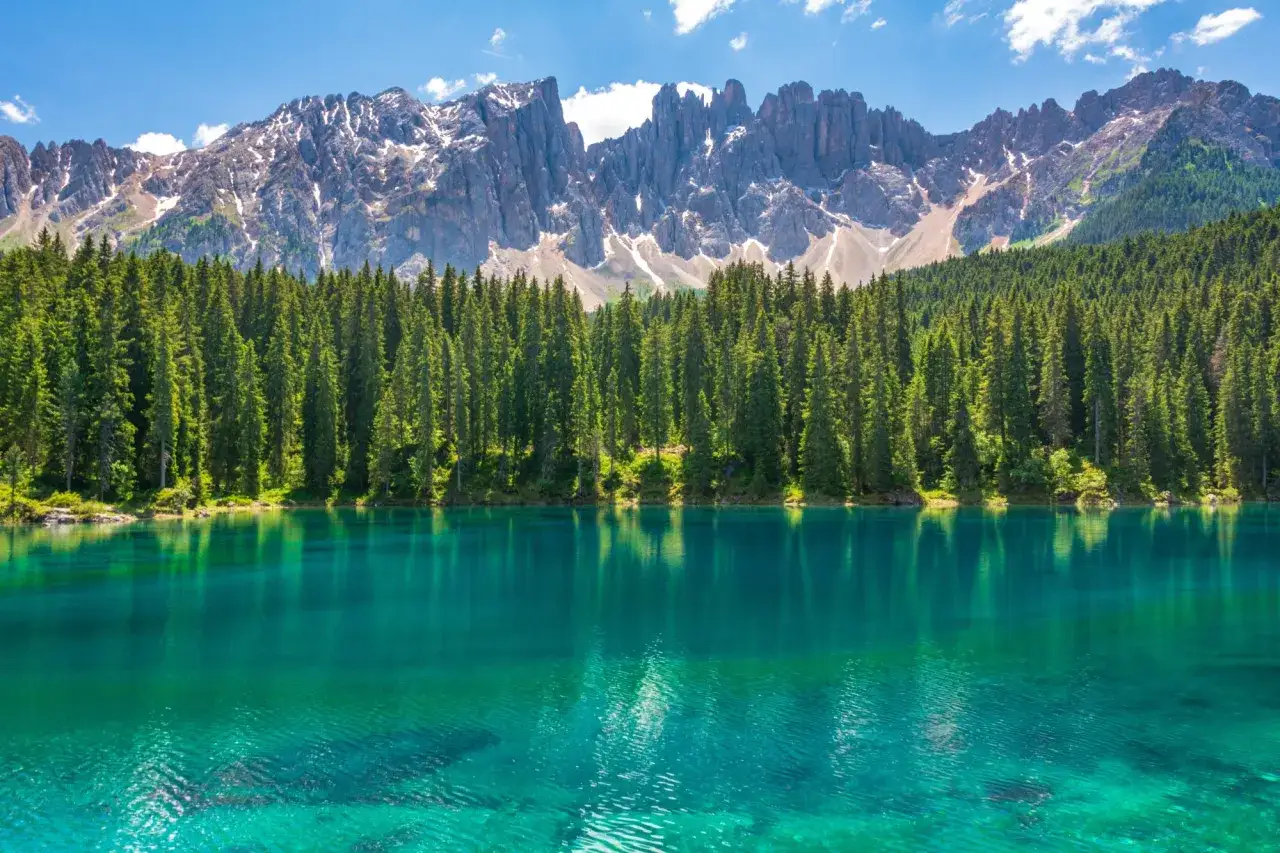Lago alpino dalle acque turchesi, circondato da una fitta foresta di abeti, con le maestose cime delle Dolomiti innevate sullo sfondo. Un paesaggio da sogno, tra Venezia e le Dolomiti.