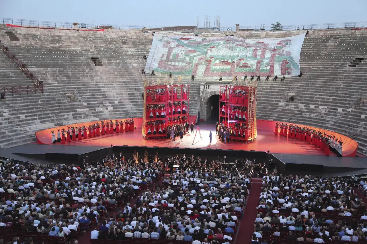 Spettacolo all'arena di Verona interno: orchestra, coro e pubblico in attesa. Un grande telo dipinto domina la scena.