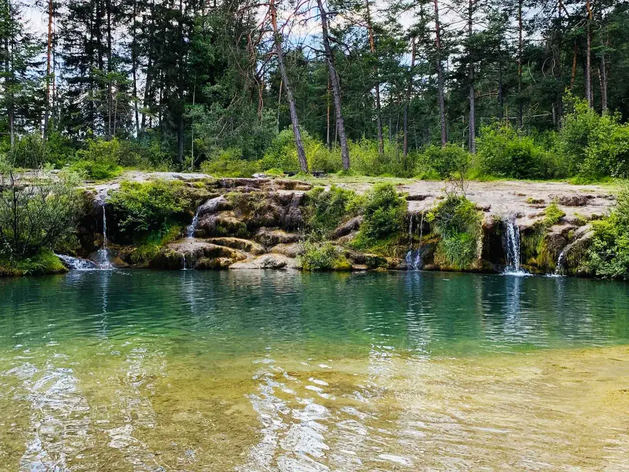 Cascate naturali nella valle di Cadore, con acqua cristallina che scorre tra rocce e vegetazione rigogliosa, circondate da un bosco di conifere.