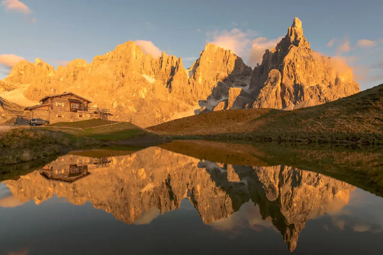 Riflesso dorato delle vette delle Dolomiti in un lago alpino, con un rifugio in legno. Perfetto per un trekking di 5 giorni.