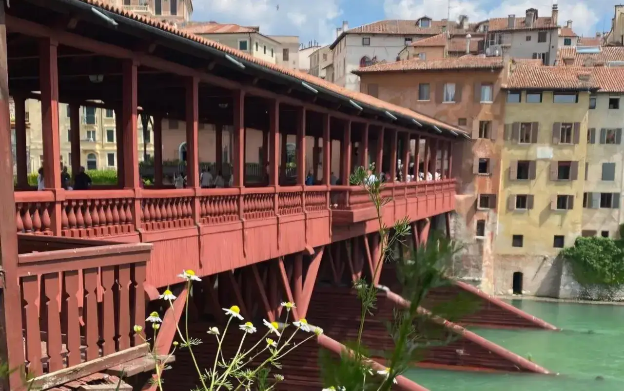 Ponte coperto rosso a Bassano del Grappa, con case colorate sullo sfondo e fiori bianchi in primo piano. Un luogo incantevole da scoprire.