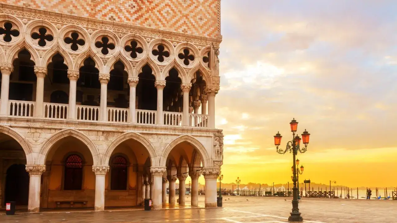 Palazzi storici di Venezia all'alba, con il sole che illumina la piazza e le architetture gotiche.