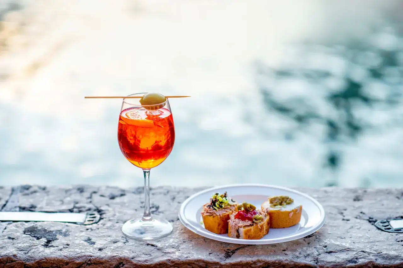 Un bicchiere di spritz venice con olive e fettina d'arancia, accompagnato da crostini, su un muretto con sfondo sfocato.