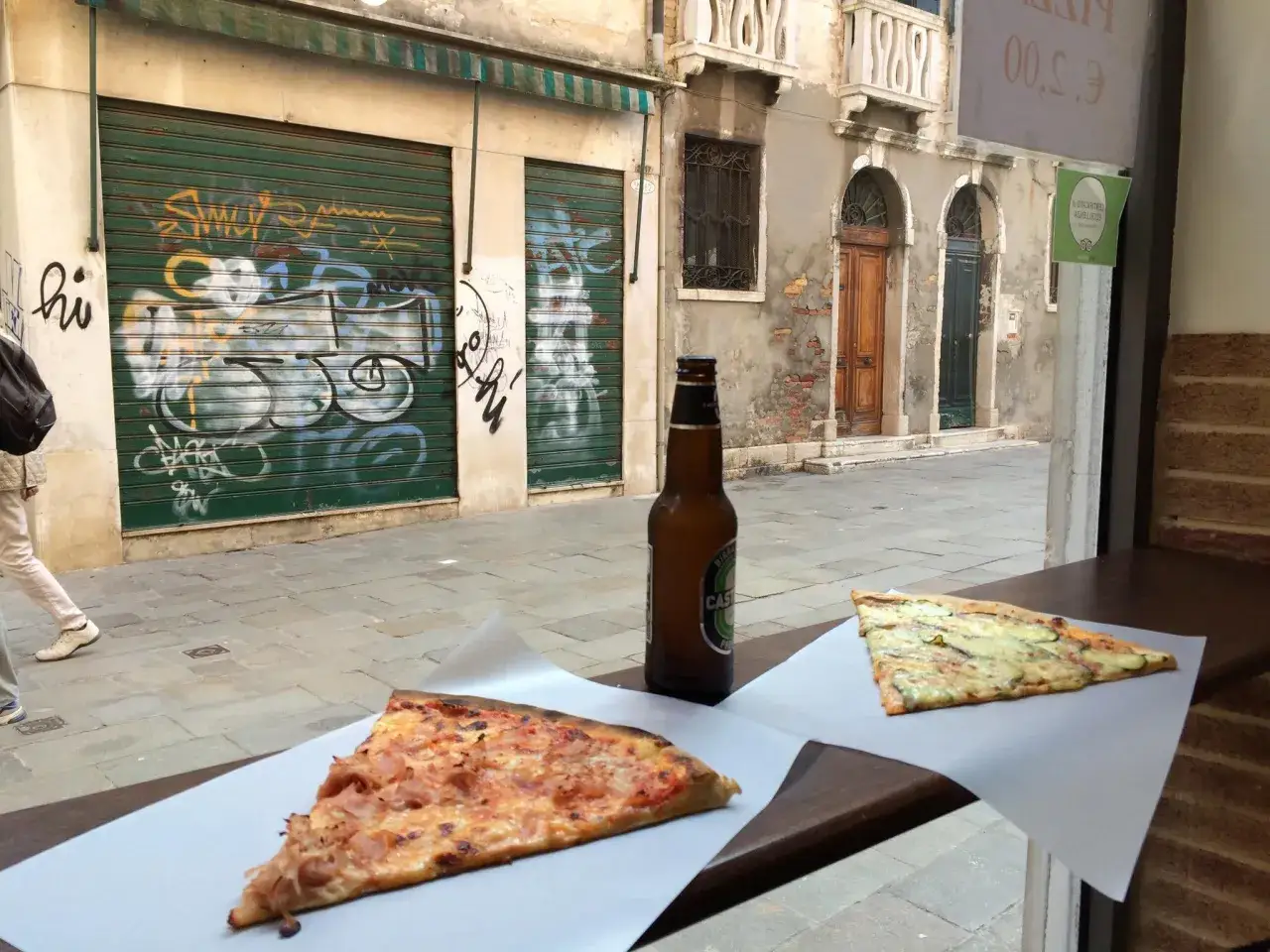 Fetta di pizza con prosciutto e zucchine, birra e un vicolo veneziano. Perfetto per trovare le migliori pizzerie Venezia.
