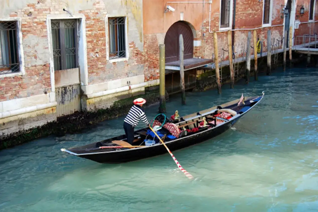 Gondoliere in abito a righe guida una delle barche tipiche veneziane lungo un canale, tra antichi palazzi.