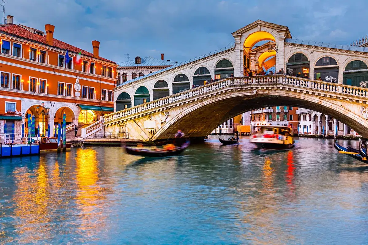 Ponte di Rialto, un'icona della storia veneziana, con gondole e vaporetti che navigano sul Canal Grande al tramonto.