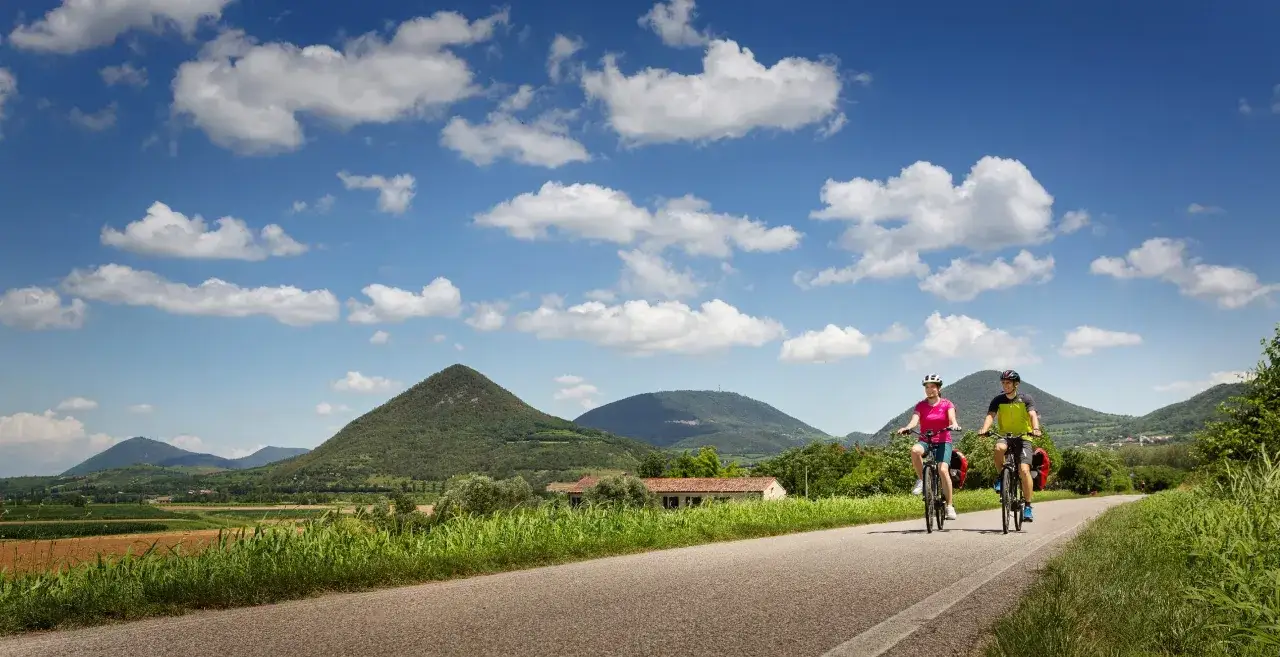 Coppia in bicicletta lungo la ciclabile dei Colli Euganei, tra verdi colline e cielo azzurro con nuvole bianche.
