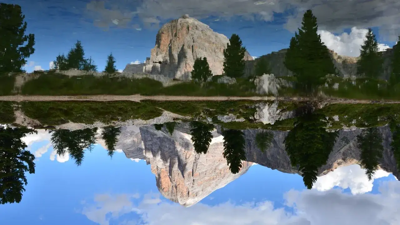 Riflesso speculare di un maestoso monte nelle Dolomiti, tra i paesi pi&ugrave; belli d'Italia. Alberi verdi e cielo azzurro completano la scena.
