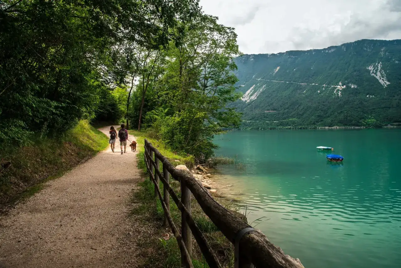 Coppia con cane passeggia lungo il sentiero del lago di Santa Croce, godendosi la natura e il panorama montano.