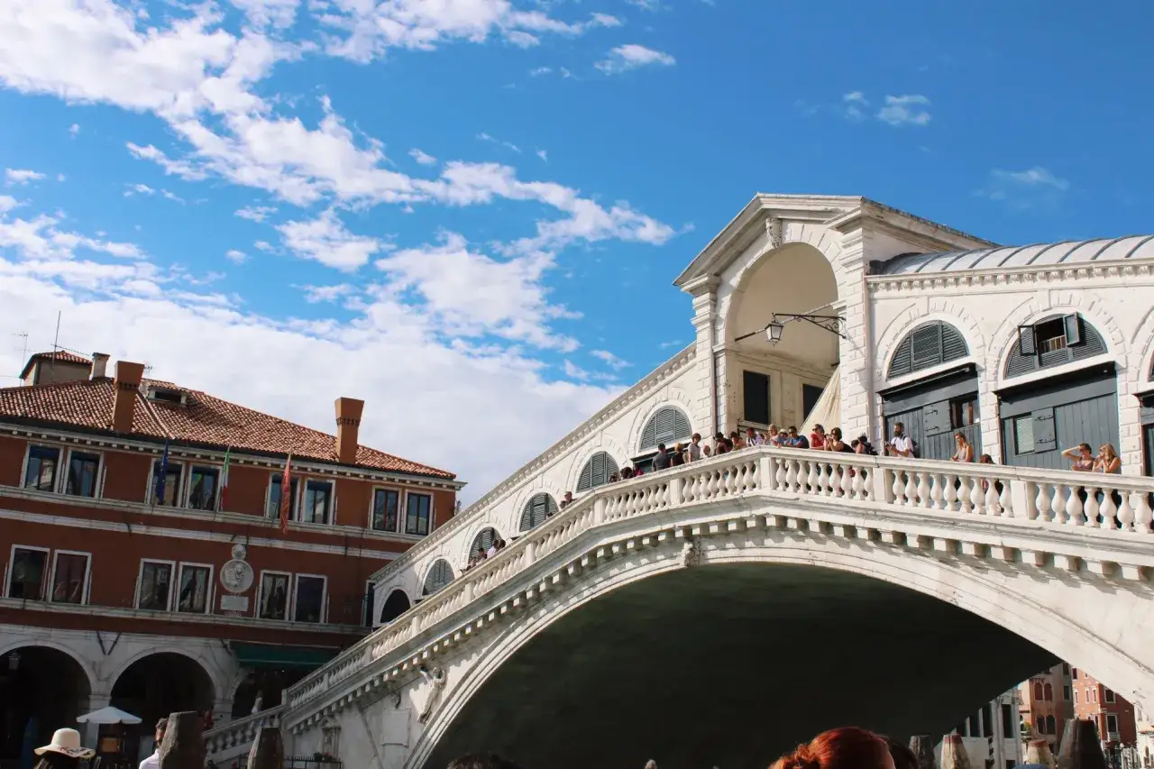 Ponte di Rialto, un'icona di cosa vedere a Venezia in un giorno. Turisti ammirano il Canal Grande sotto un cielo azzurro con nuvole bianche.