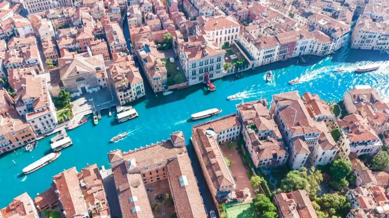 Vista aerea del **canale di Venezia** con gondole e vaporetti che navigano tra palazzi storici dai tetti rossi.